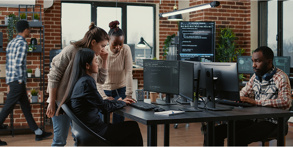 Diverse team in a modern tech office: woman codes on dual screens, two colleagues discuss. Man codes independently at another desk.